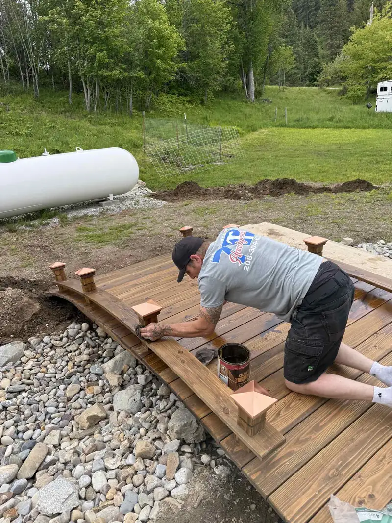 7B Handyman crew member in a 7B Handyman shirt staining the railing and copper-capped posts of a wooden garden bridge