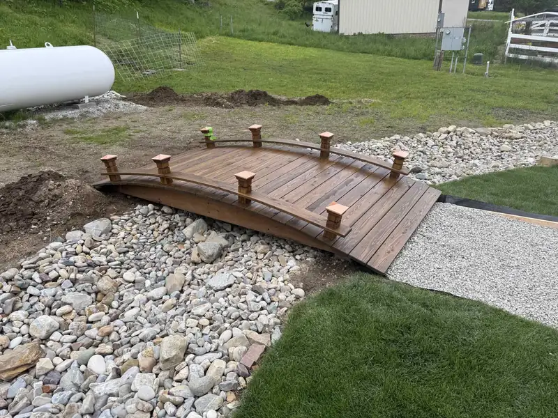 Finished wooden garden bridge over a rock-lined dry creek bed, built and stained by 7B Handyman in Sandpoint, Idaho