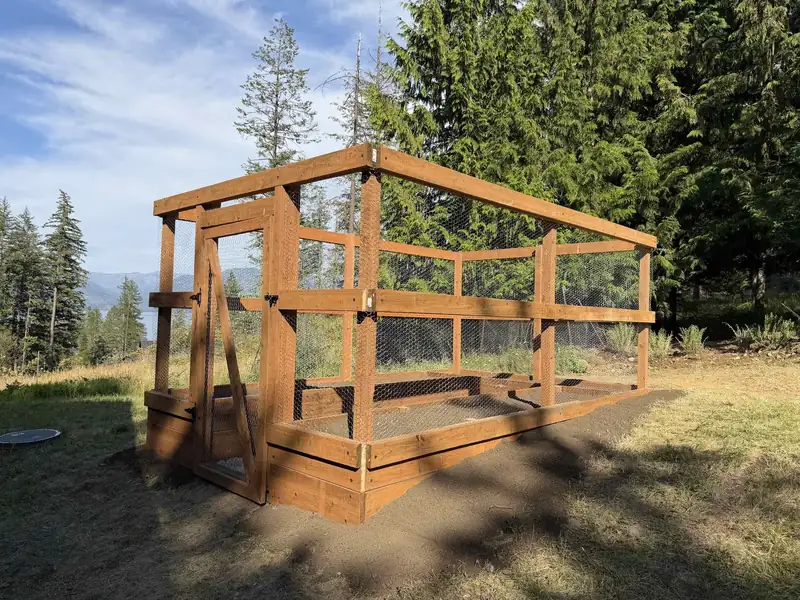 Wood-frame garden and coop enclosure under construction with a lake and mountain view in the background